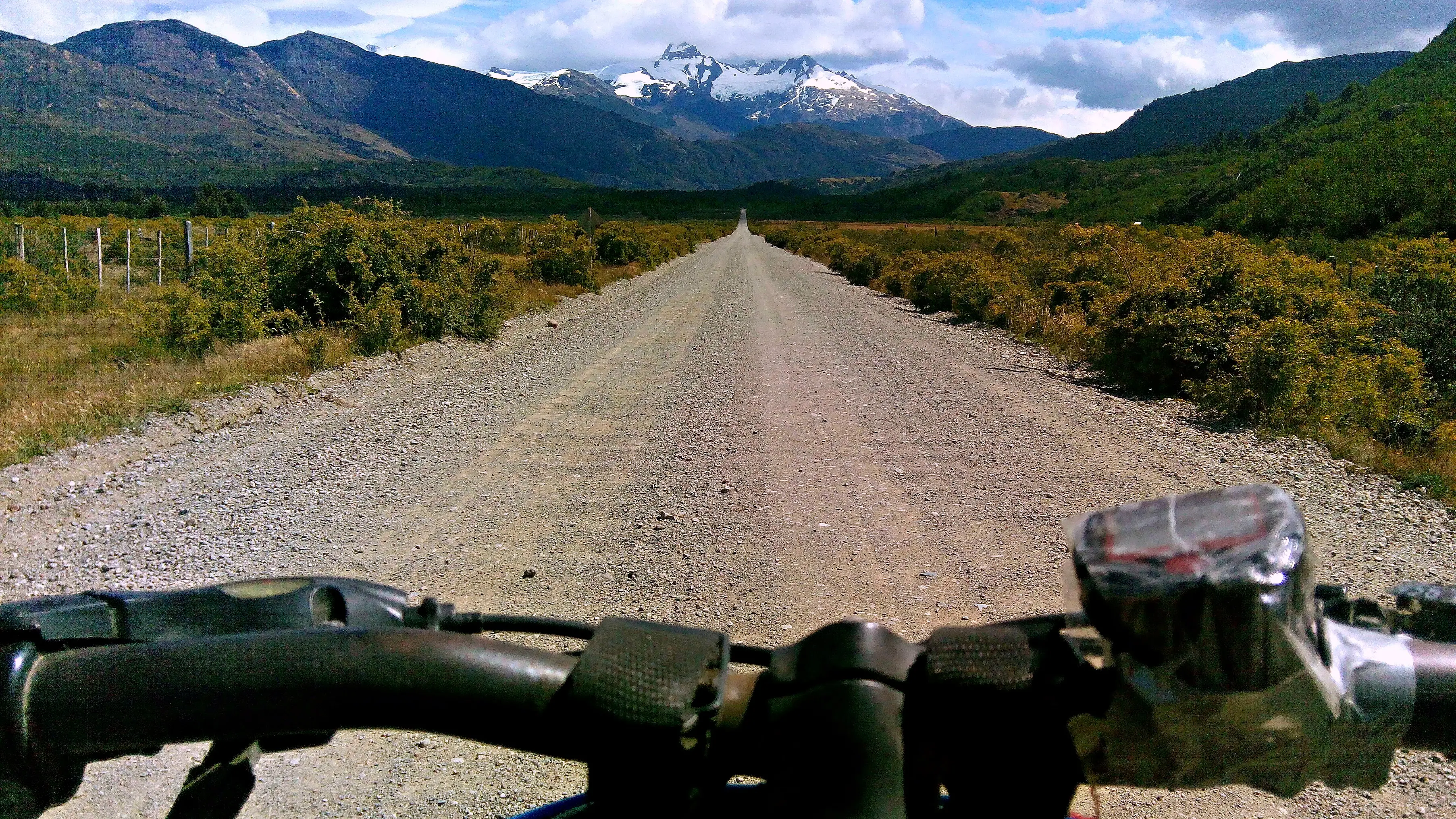 Vista desde el manubrio de una bicicleta en un camino de ripio de la Carretera Austral, con montañas nevadas al fondo