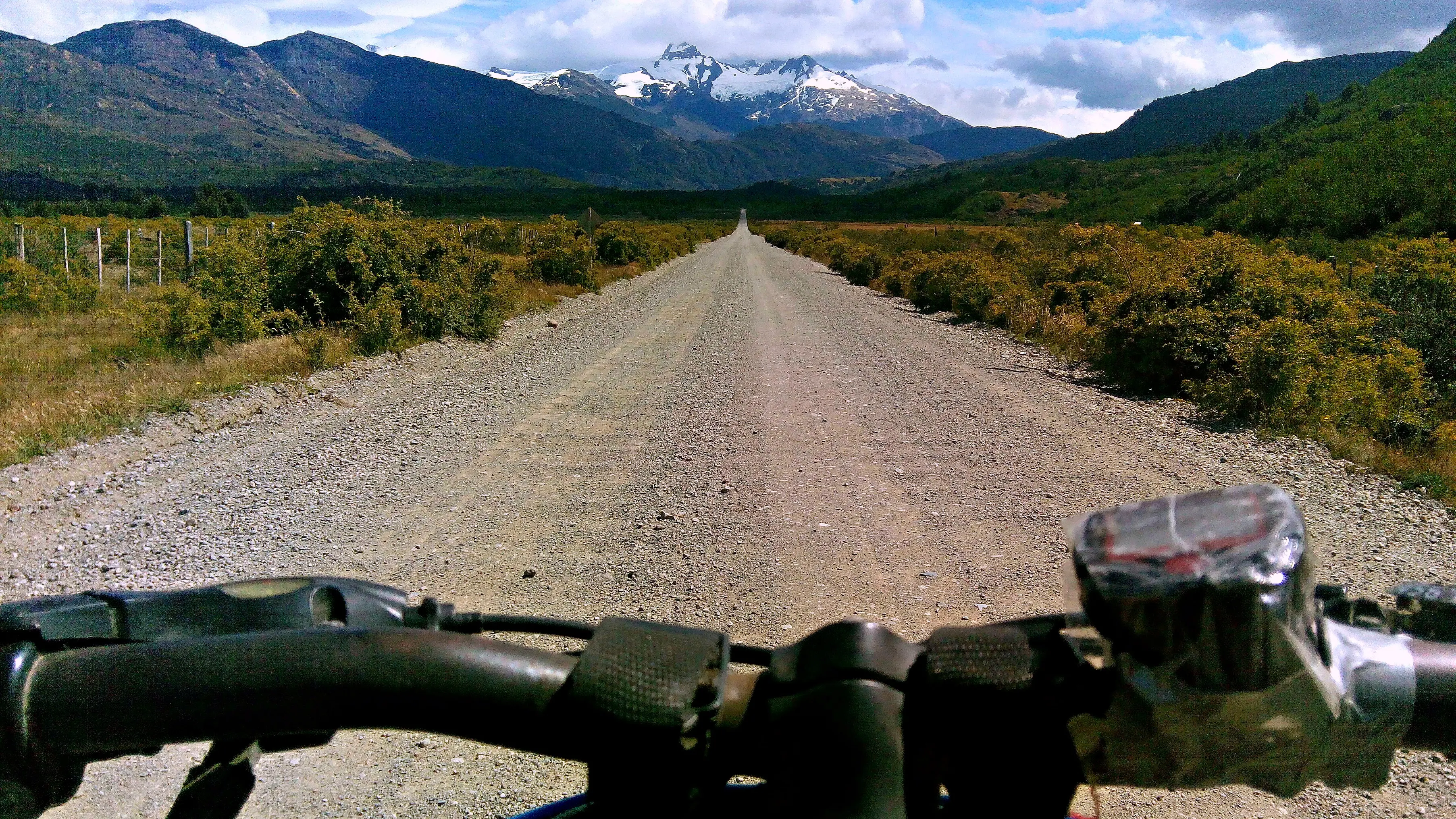 Vista desde el manubrio de una bicicleta en un camino de ripio de la Carretera Austral, con montañas nevadas al fondo